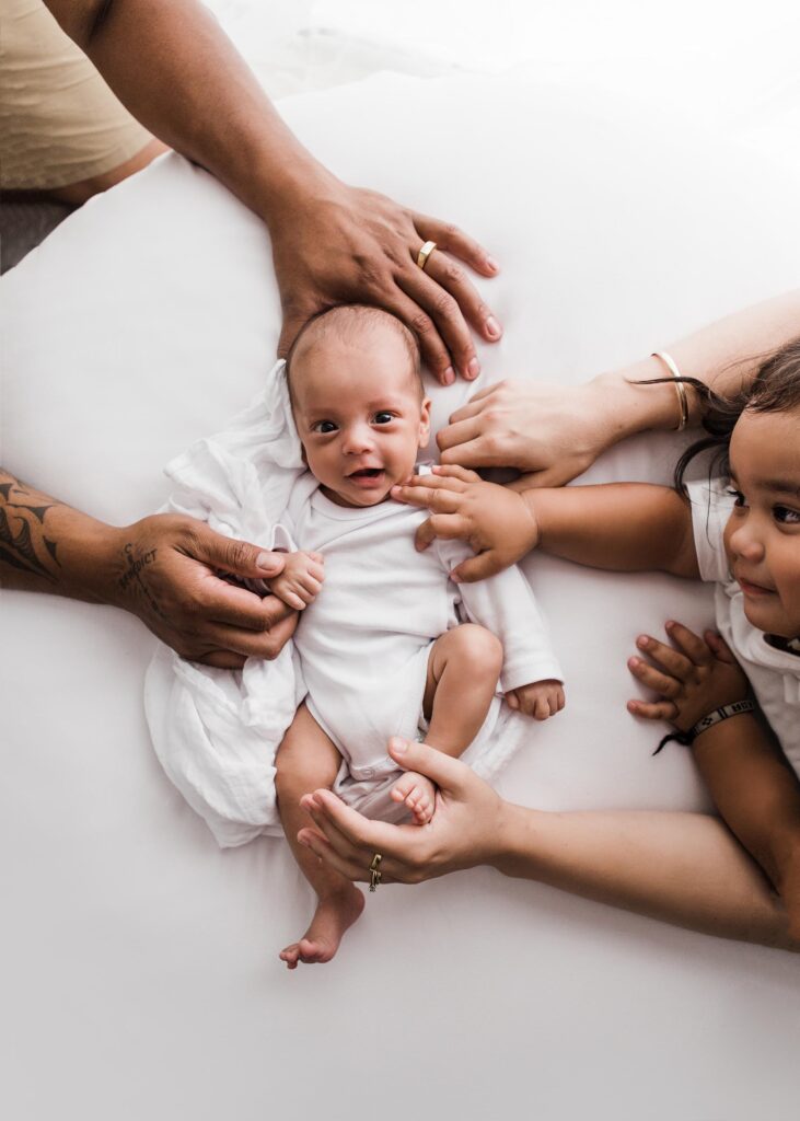 Newborn baby surrounded by the hands of his family memebers. Baby is smiling and looking straight into the camera. 