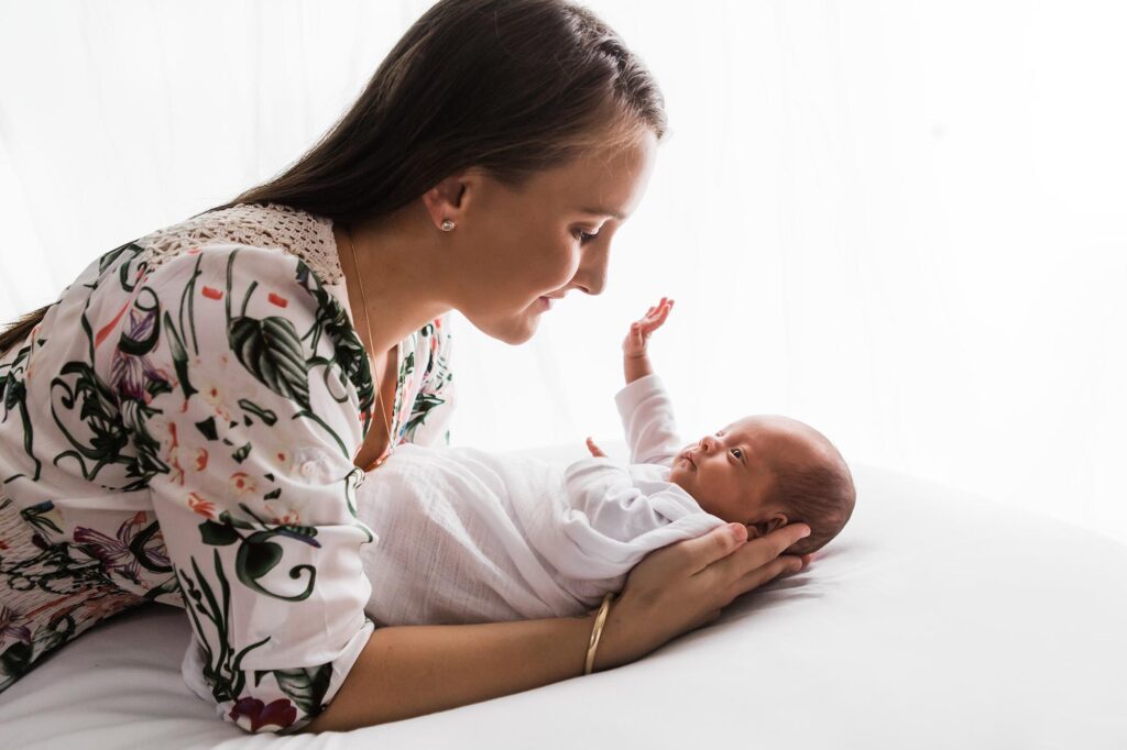 Newborn baby being held by his mother who is leaning over and looking into his eyes. Baby has his hand in the air stretching out towards his mother's face. 