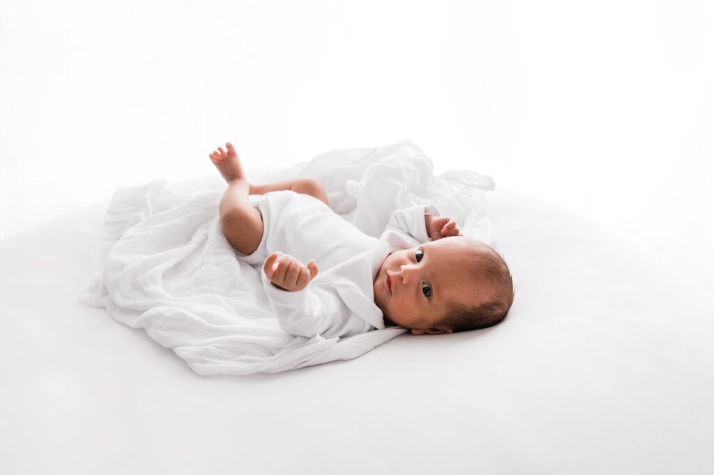 Newborn baby wearing white onesie awake looking into camera during photoshoot.