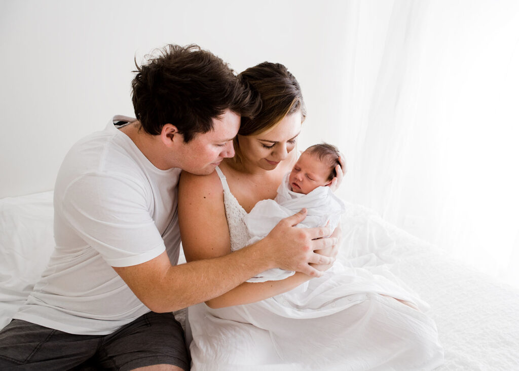 Mother holding her newborn baby while father wraps arms around mother and baby during thier newborn photo shoot.