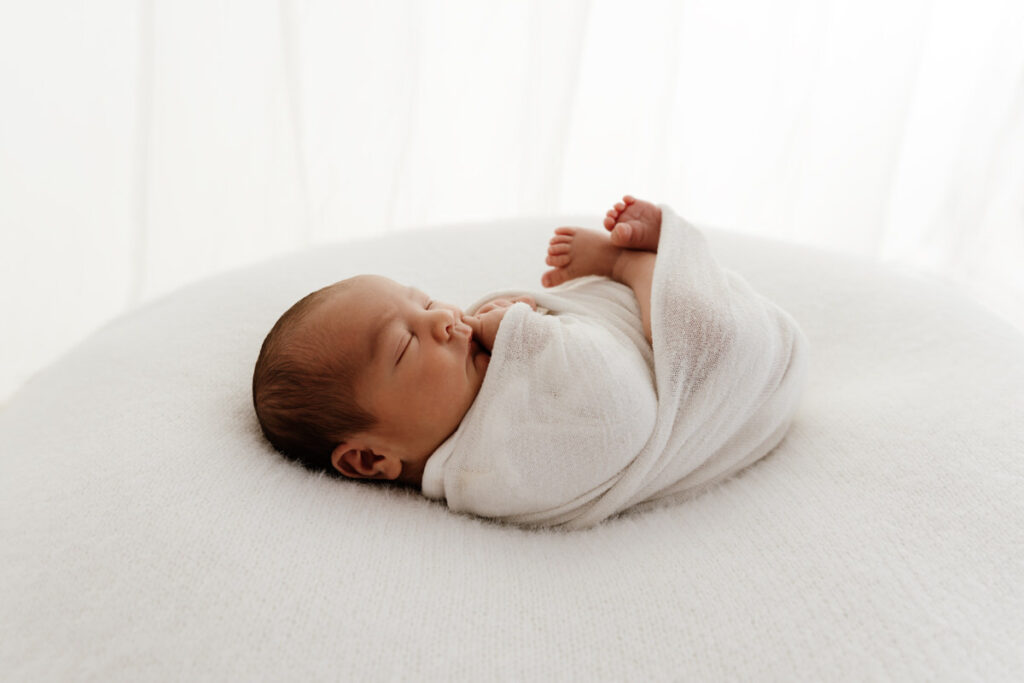 Newborn baby swaddles in white wrap. Baby sleeps during his newborn photo shoot.