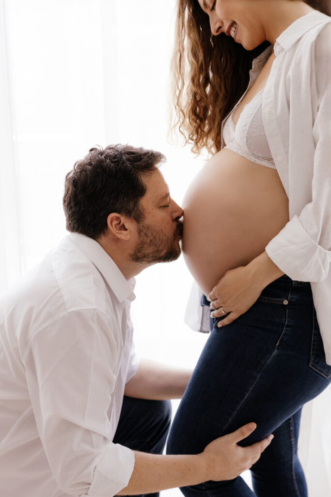 In the maternity photoshoot, the husband is kissing his wife's baby bump, and the wife is smiling in Abu Dhabi