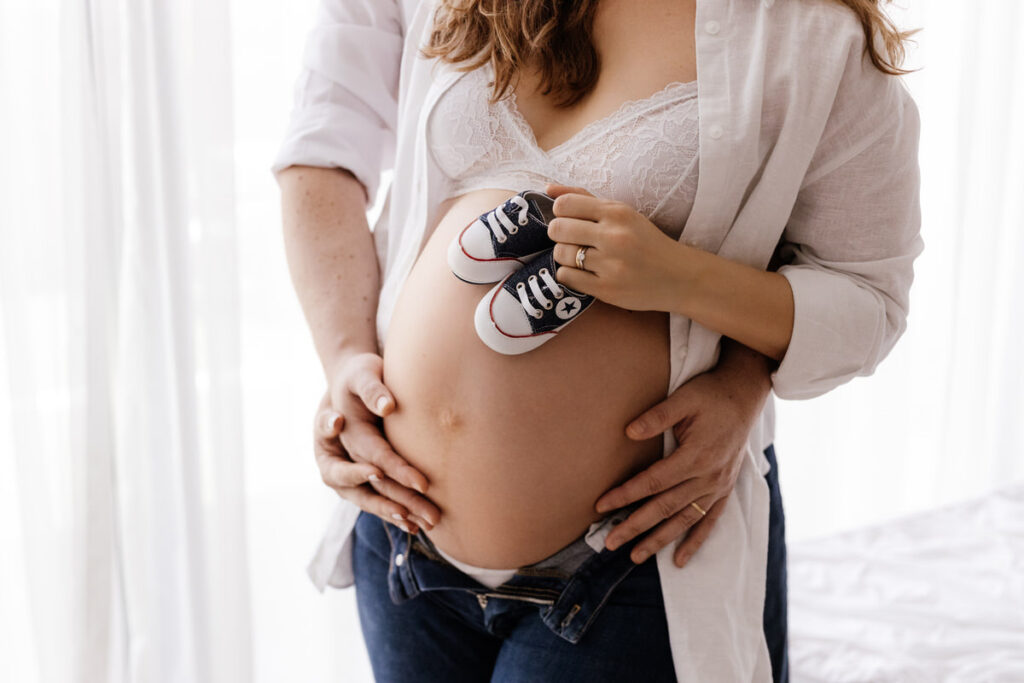 The husband is standing behind his wife during a maternity photo shoot, gently holding her baby bump from behind with his hands, while the wife is touching the baby bump with one hand, showing the newborn baby's shoes. The scene is set in Abu Dhabi